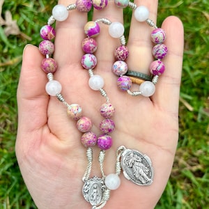 May include: A silver and white rosary with pink and white beads. The beads are made of a speckled stone. The rosary has a silver charm with a picture of the Virgin Mary and a silver charm with a picture of Saint Anthony.