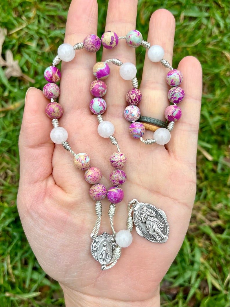 May include: A silver and white rosary with pink and white beads. The beads are made of a speckled stone. The rosary has a silver charm with a picture of the Virgin Mary and a silver charm with a picture of Saint Anthony.
