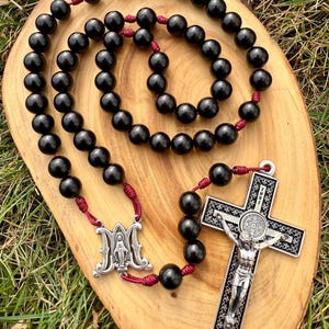 May include: A black rosary with dark red cord and silver crucifix and centerpiece. The rosary beads are round and smooth. The crucifix features a detailed depiction of Jesus Christ. The centerpiece has an image of the Virgin Mary.