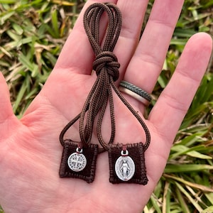 May include: Two small brown fabric pouches with silver charms hanging from brown cords. The charms are a silver oval with a woman's image and a silver cross with four smaller crosses.
