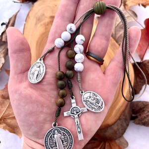 May include: A green, brown, and white beaded rosary with a silver crucifix, a silver medal of Saint Michael, a silver medal of the Virgin Mary, and a silver medal of Saint Anthony. The rosary is held in a hand.