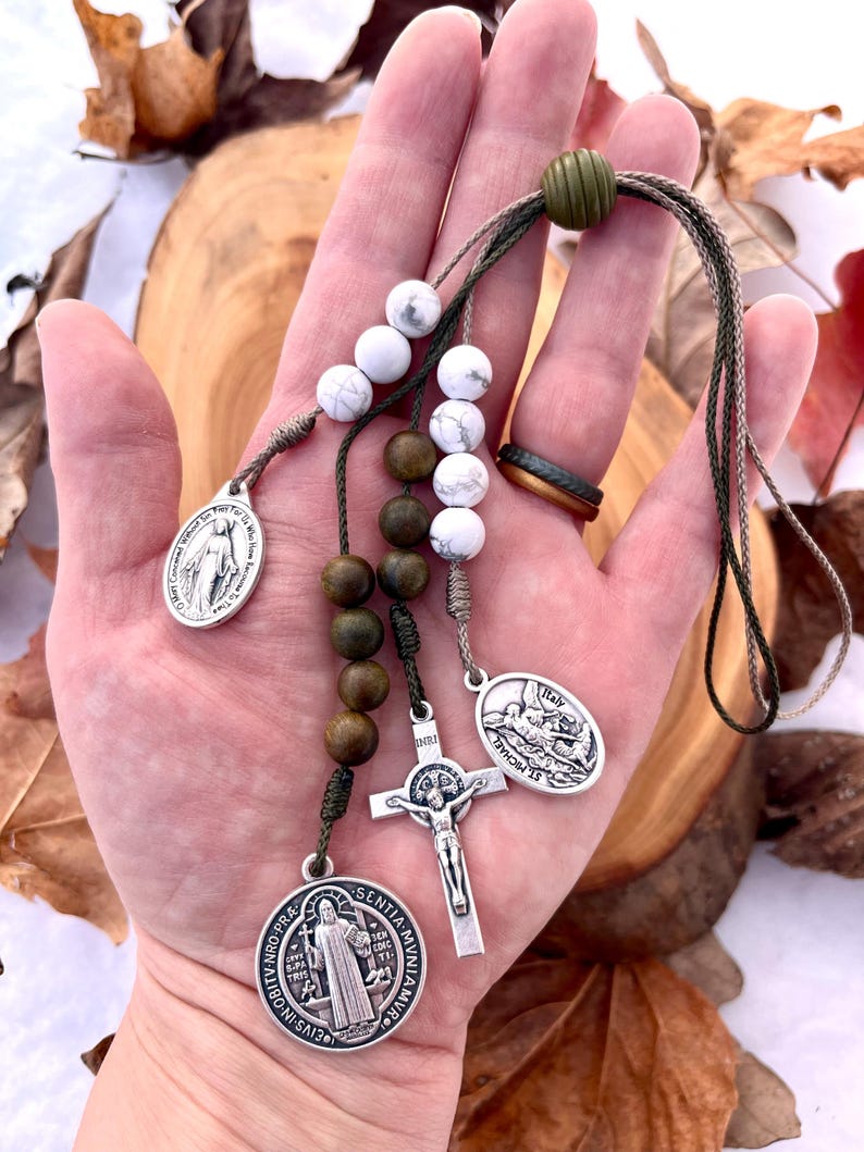 May include: A green, brown, and white beaded rosary with a silver crucifix, a silver medal of Saint Michael, a silver medal of the Virgin Mary, and a silver medal of Saint Anthony. The rosary is held in a hand.