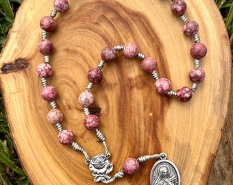 May include: A rosary with pink and white speckled beads strung on silver wire. The rosary features a silver rose centerpiece and a silver pendant with a religious figure. The beads are arranged on a wooden surface.