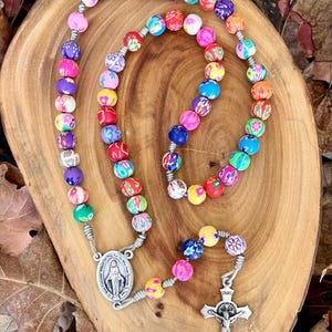 May include: A colourful rosary with floral patterned beads, a silver metal pendant featuring the Virgin Mary, and a silver metal crucifix with the inscription "INRI".