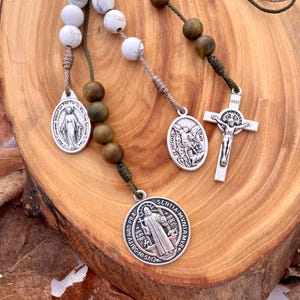 May include: A green and brown rosary with white beads and silver charms. The charms feature images of the Virgin Mary, St. Michael, and a crucifix with the inscription "INRI".