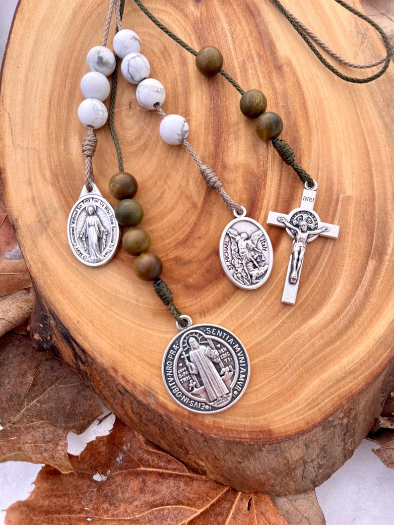 May include: A green and brown rosary with white beads and silver charms. The charms feature images of the Virgin Mary, St. Michael, and a crucifix with the inscription "INRI".
