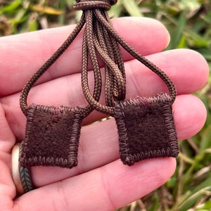 May include: Two brown suede squares with stitched edges are attached to a brown cord. The cord is tied in a knot.