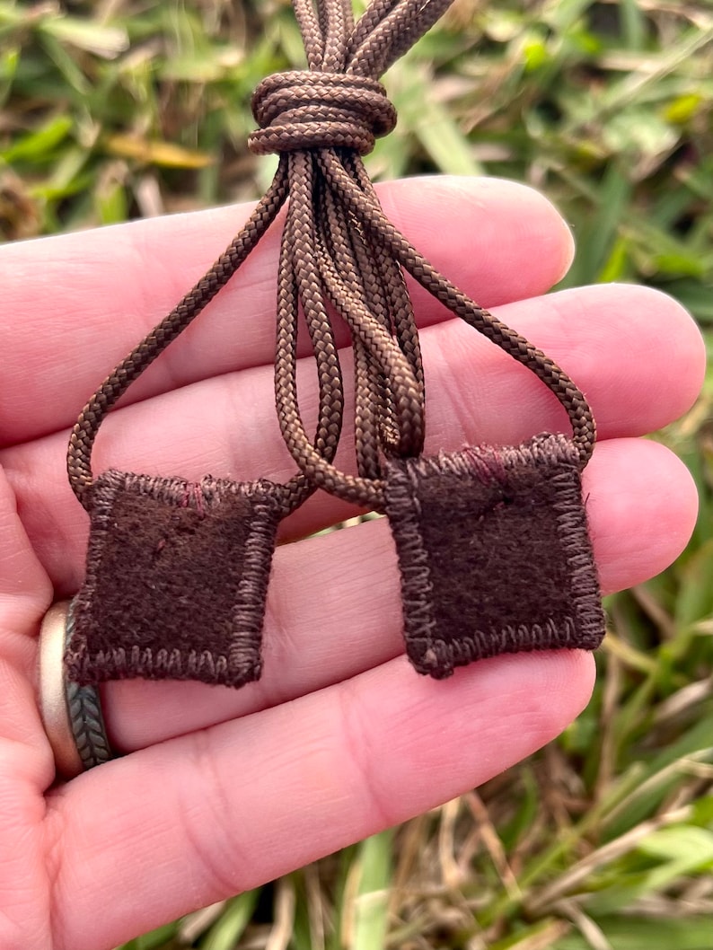 May include: Two brown suede squares with stitched edges are attached to a brown cord. The cord is tied in a knot.