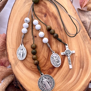 May include: A green, brown, and white beaded rosary with a silver pendant featuring the image of Saint Michael, a silver pendant featuring the image of the Virgin Mary, and a silver crucifix with the inscription "INRI".