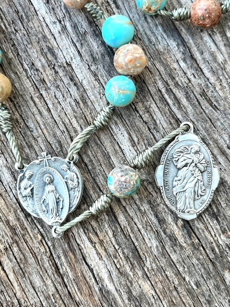 May include: A silver rosary with turquoise and brown stone beads. The rosary features two silver medallions with religious imagery. One medallion depicts the Virgin Mary and the other depicts Our Lady Untier of Knots.