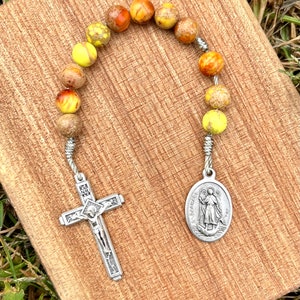 May include: A yellow, orange, and brown stone rosary with a silver cross and a silver oval charm featuring a saint. The charm has the text "St. Raphael" on it.