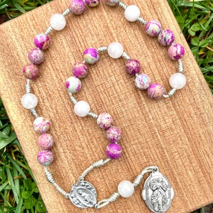 May include: A silver rosary with pink and white beads. The beads are made of a marbled stone. The rosary has a silver charm with a religious image.