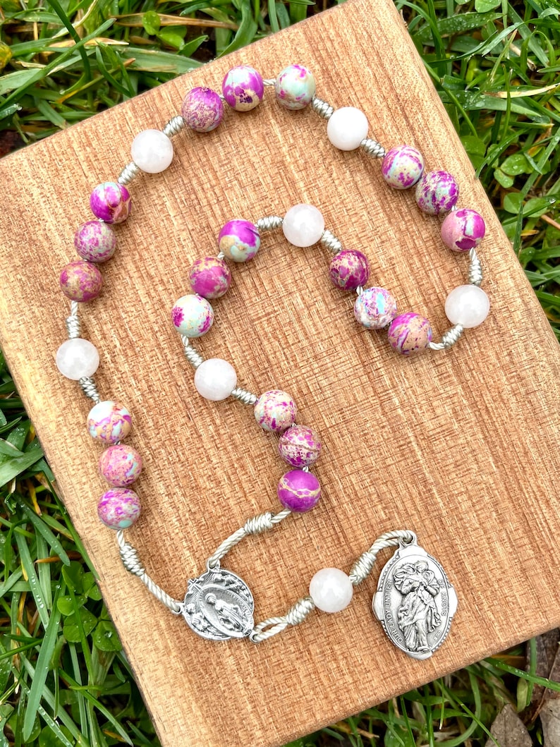 May include: A silver rosary with pink and white beads. The beads are made of a marbled stone. The rosary has a silver charm with a religious image.