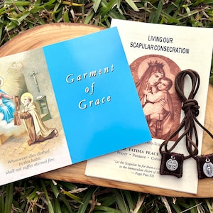 May include: A brown scapular with two medallions, one with a cross and the other with the Virgin Mary. The scapular is on a wooden surface with two pamphlets, one titled "Garment of Grace" and the other titled "Living Our Scapular Consecration".