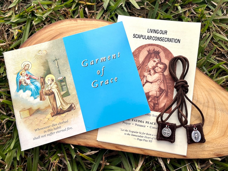 May include: A brown scapular with two medallions, one with a cross and the other with the Virgin Mary. The scapular is on a wooden surface with two pamphlets, one titled "Garment of Grace" and the other titled "Living Our Scapular Consecration".