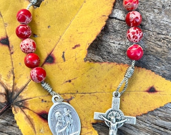 Op de afbeelding: Rode en witte gevlekte stenen rozenkrans kralen met een zilveren kruisbeeld en een zilveren medaille van de Heilige Judas Thaddeus. De rozenkrans ligt op een geel blad.