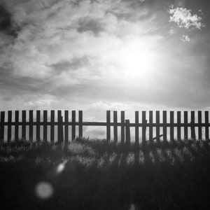 May include: A black and white photo of a wooden fence with a gap in the middle. The fence is in front of a field of grass and a cloudy sky. The sun is shining through the clouds.