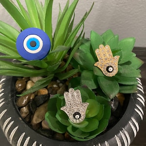 Puede incluir: Tres amuletos de mano de hamsa con un ojo turco en el centro. Las manos de hamsa están decoradas con diamantes de imitación. Una mano de hamsa es dorada, una es plateada y una es azul.