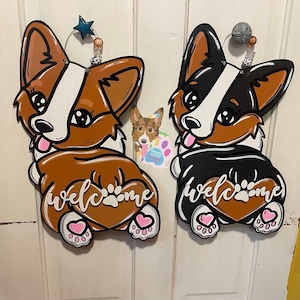 Puede incluir: Dos letreros de bienvenida de madera con forma de corgi. El corgi de la izquierda es marr&oacute;n y blanco con la palabra "bienvenido" escrita en forma de coraz&oacute;n. El corgi de la derecha es negro y blanco con la palabra "bienvenido" escrita en forma de coraz&oacute;n.