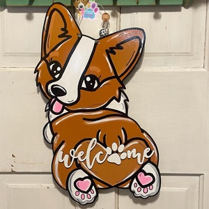 Puede incluir: Un colgador de puerta de madera con forma de perro corgi marr&oacute;n y blanco con corazones rosas en las patas. El perro est&aacute; sentado de espaldas al espectador y tiene una cola en forma de coraz&oacute;n con la palabra "bienvenido" escrita en ella.