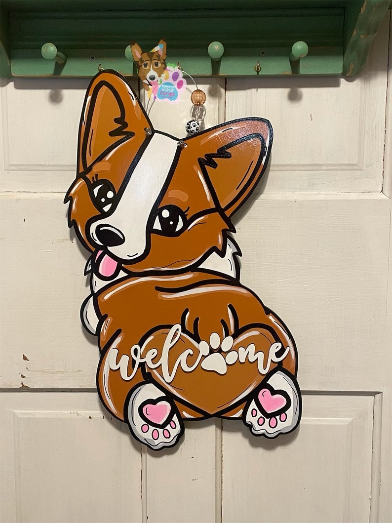 Puede incluir: Un colgador de puerta de madera con forma de perro corgi marr&oacute;n y blanco con corazones rosas en las patas. El perro est&aacute; sentado de espaldas al espectador y tiene una cola en forma de coraz&oacute;n con la palabra "bienvenido" escrita en ella.