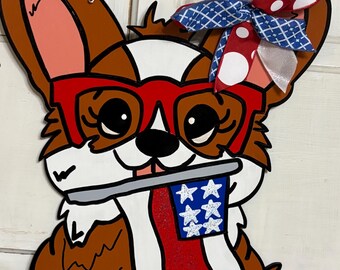 May include: Wooden corgi dog door hanger with red glasses, holding a patriotic flag. The dog is brown, white, and black, with pink accents. A red, white, and blue bow adorns the top, and a beaded wire hanger is attached.