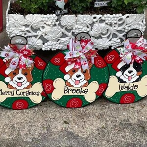 May include: Three green and red Christmas ornament-shaped wooden signs with dog illustrations and names. The signs read "Merry Corgmas", "Brooke", and "Waldo".