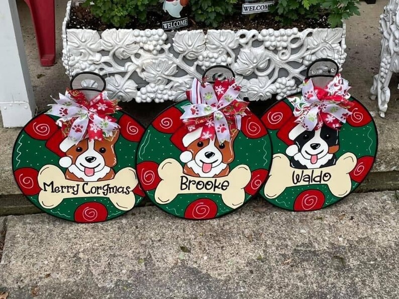 May include: Three green and red Christmas ornament-shaped wooden signs with dog illustrations and names. The signs read "Merry Corgmas", "Brooke", and "Waldo".