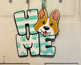 May include: Wooden "HOME" sign with a Corgi dog design. The letters are white with teal accents, including stripes, paw prints, and polka dots. The Corgi has a brown and white coat with pink ears and a happy expression.