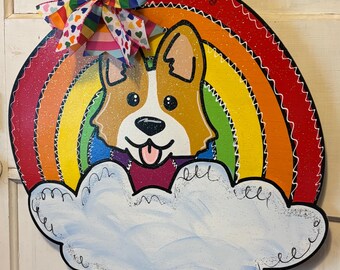 May include: A round wooden door hanger with a rainbow and a cartoon corgi dog. The corgi is wearing a purple sweater and has a pink tongue sticking out. The rainbow is made of red, orange, yellow, green, blue, indigo, and violet. The door hanger is decorated with white stitching and glitter.