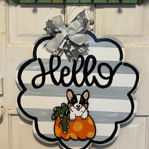 May include: A gray and white striped door hanger with the word "Hello" in black script. A cartoon corgi dog is sitting in a pumpkin with orange and brown swirls. A gray and white bow with bone print is attached to the top.