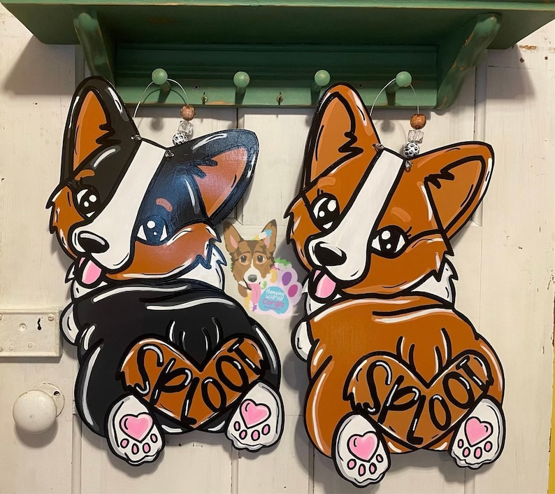 May include: Two wooden cutouts of corgis in a "sploot" pose. The corgis are painted in black, white, brown, and orange. The corgis have pink hearts on their backs with the word "sploot" written on them.