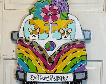 May include: A colorful wooden cutout of a vintage van with a peace sign and rainbow stripes. The van has two corgi dogs inside and flowers on top. The text "Don't Worry... Be Happy" is on the front of the van.
