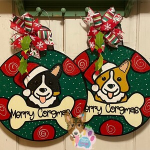 May include: Two Christmas ornaments featuring a black and white and a brown and white corgi dog wearing Santa hats. The ornaments are green with red and white swirls and the text "Merry Corgmas" on a bone.
