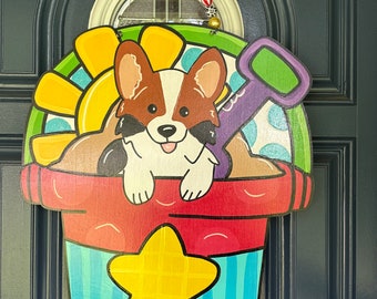 May include: A colorful wooden door hanger featuring a cartoon corgi dog peeking out of a red and blue bucket with a yellow star. The bucket is surrounded by a yellow sunflower and a purple shovel.
