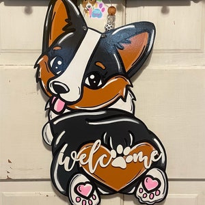 Puede incluir: Un colgador de puerta de madera con forma de perro corgi blanco y negro con orejas marrones y un coraz&oacute;n marr&oacute;n en el vientre. El coraz&oacute;n tiene una huella de pata blanca y la palabra "welcome" escrita en letras blancas cursivas. El perro tiene corazones rosas en sus patas.