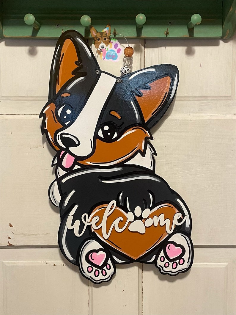 Puede incluir: Un colgador de puerta de madera con forma de perro corgi blanco y negro con orejas marrones y un coraz&oacute;n marr&oacute;n en el vientre. El coraz&oacute;n tiene una huella de pata blanca y la palabra "welcome" escrita en letras blancas cursivas. El perro tiene corazones rosas en sus patas.