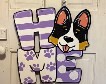 May include: A wooden "Home" sign with a purple, white, and black dog illustration. The letters are painted in purple and white stripes, paw prints, and white polka dots.