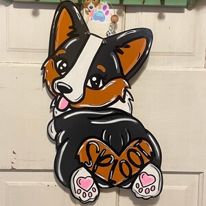 May include: A wooden door hanger shaped like a black and white corgi dog with brown ears and a brown heart on its back that says "Spoot". The dog has pink hearts on its paws and is smiling.
