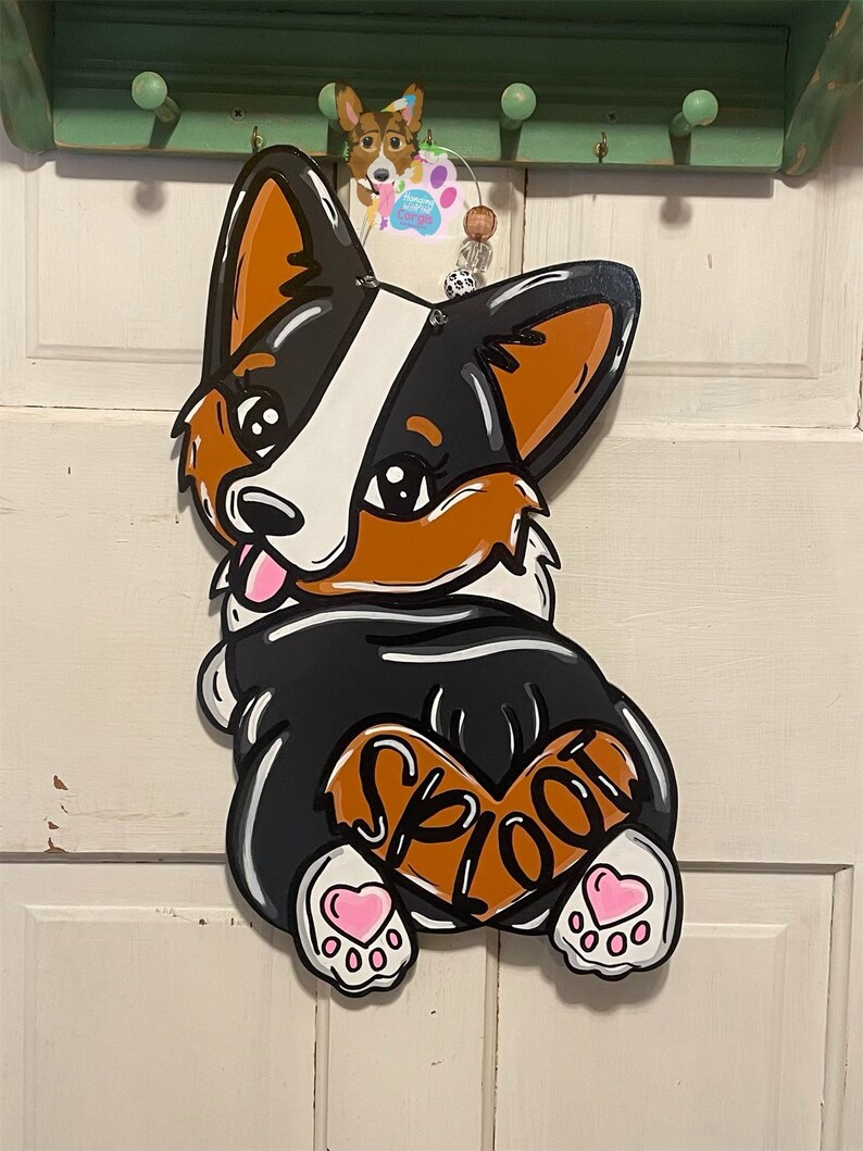 May include: A wooden door hanger shaped like a black and white corgi dog with brown ears and a brown heart on its back that says "Spoot". The dog has pink hearts on its paws and is smiling.