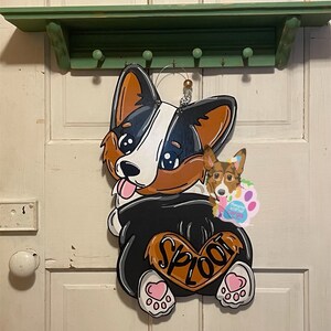 May include: A wooden door hanger shaped like a corgi dog with a black and white body, brown ears, and a pink heart on its rear end. The dog is wearing a black shirt with the word "SPLOOT" written on it. The dog is holding a smaller corgi dog in its mouth.
