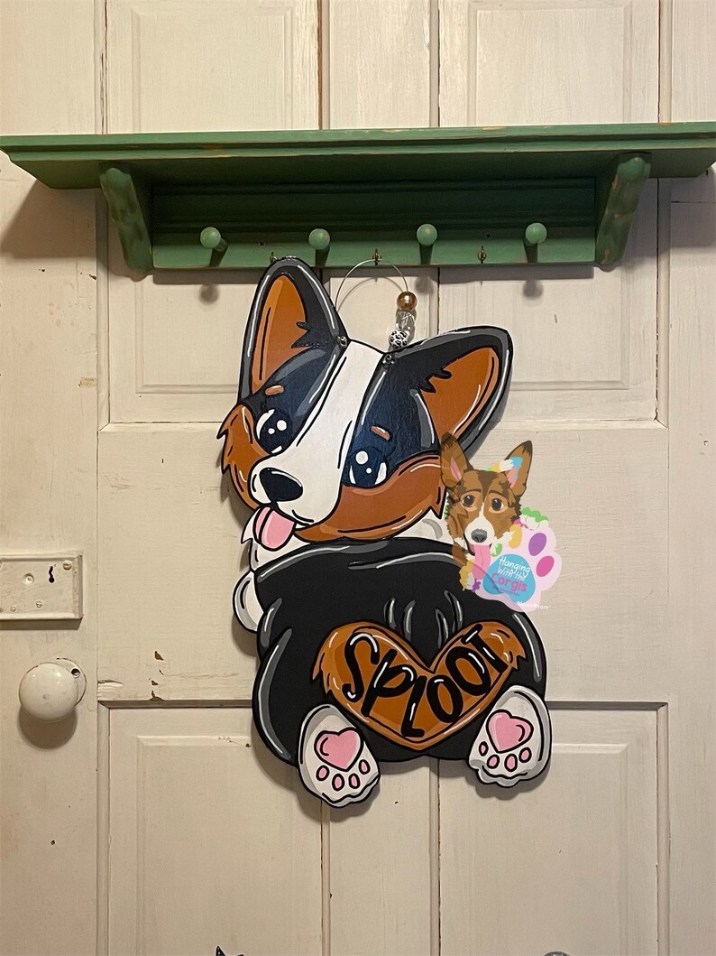 May include: A wooden door hanger shaped like a corgi dog with a black and white body, brown ears, and a pink heart on its rear end. The dog is wearing a black shirt with the word "SPLOOT" written on it. The dog is holding a smaller corgi dog in its mouth.