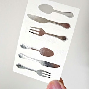 May include: A sheet of silver-colored cutlery stickers. The stickers include spoons, knives, and forks. The utensils have decorative handles and are arranged on a white background. The stickers are held by a person's hand.