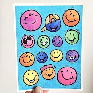 May include: A rectangular greeting card featuring a bright blue background covered in various colorful smiley face illustrations. The faces include different expressions, colors, and accessories like a baseball cap, sunglasses, and a small pink hat.