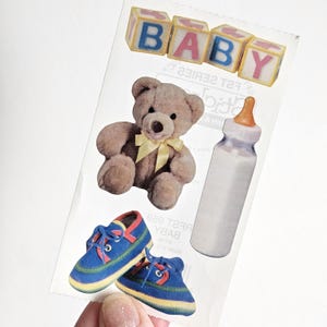 May include: A sticker sheet featuring baby-themed images. Includes blocks spelling "BABY", a teddy bear with a yellow bow, a baby bottle, and blue sneakers with red and yellow accents.