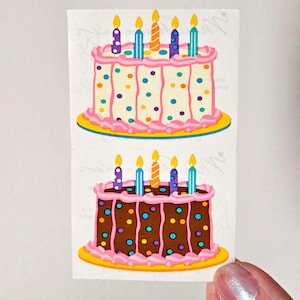 May include: Two birthday cake stickers. The top cake is white with pink frosting, colorful polka dots, and six candles. The bottom cake is brown with pink frosting, colorful polka dots, and six candles. Both cakes sit on yellow plates.