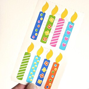May include: A sheet of colorful birthday candle stickers. Each candle has a yellow flame and a different design on the body, including polka dots, flowers, stripes, and hearts. The candles are in shades of blue, green, pink, and orange.