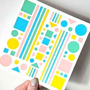 May include: A white square with a colorful geometric design. The design features shapes like squares, circles, triangles, and stripes in pastel colors such as pink, blue, yellow, and green. The artwork is held by a hand with a glittery nail.
