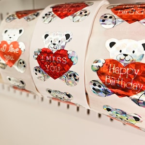 May include: Rolls of stickers featuring white teddy bears holding red heart-shaped balloons. The hearts have messages like "I love you," "I miss you," and "Happy Birthday." The stickers have a holographic, iridescent effect.