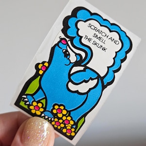 May include: A rectangular sticker featuring a cartoon skunk in blue with a white speech bubble. The text "SCRATCH AND SMELL THE SKUNK" is in the speech bubble. The skunk is surrounded by yellow and pink flowers and green leaves.